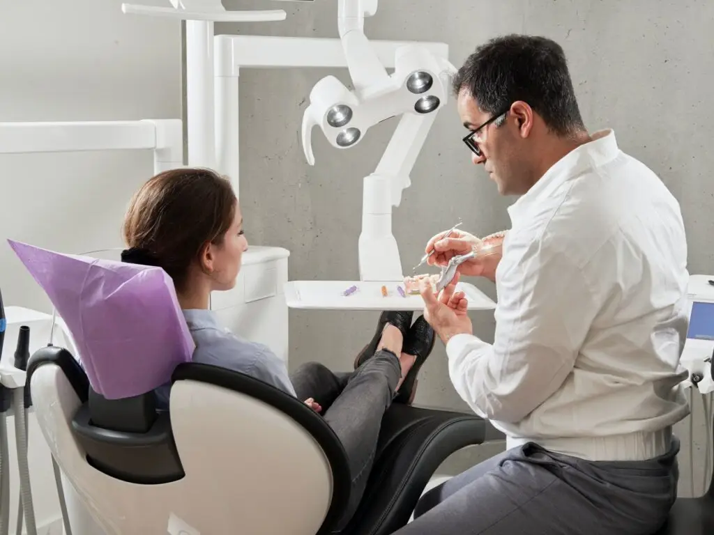 Dentysta objaśnia model protetyczny siedzącemu pacjentowi w nowoczesnej klinice. Pacjent słucha uważnie, gdy dentysta gestykuluje do modelu, otoczony białym sprzętem dentystycznym i jasnymi światłami.