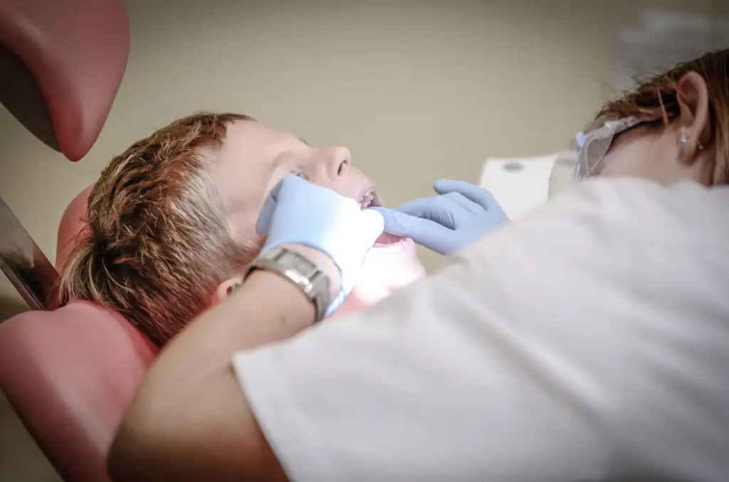 Dentysta w rękawiczkach bada jamę ustną małego chłopca podczas wizyty u dentysty, który leży na fotelu dentystycznym w jasnym świetle.