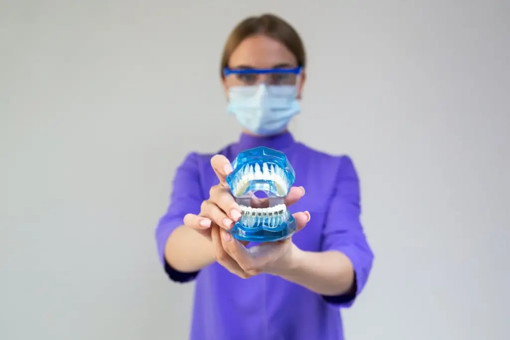 Dentysta w masce i okularach ochronnych trzyma model dentystyczny z aparatem ortodontycznym w kierunku kamery, podkreślając znaczenie ortodoncji w zapobieganiu chorobie dziąseł, na prostym tle.