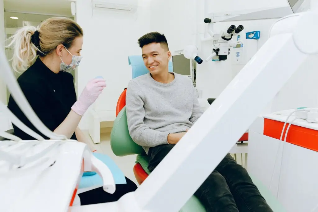 Dentysta w rękawiczkach i masce rozmawia z uśmiechniętym pacjentem siedzącym na fotelu dentystycznym w jasnej, nowoczesnej klinice, pomagając złagodzić strach przed wizytą u stomatologa. W pomieszczeniu widoczny jest sprzęt dentystyczny.