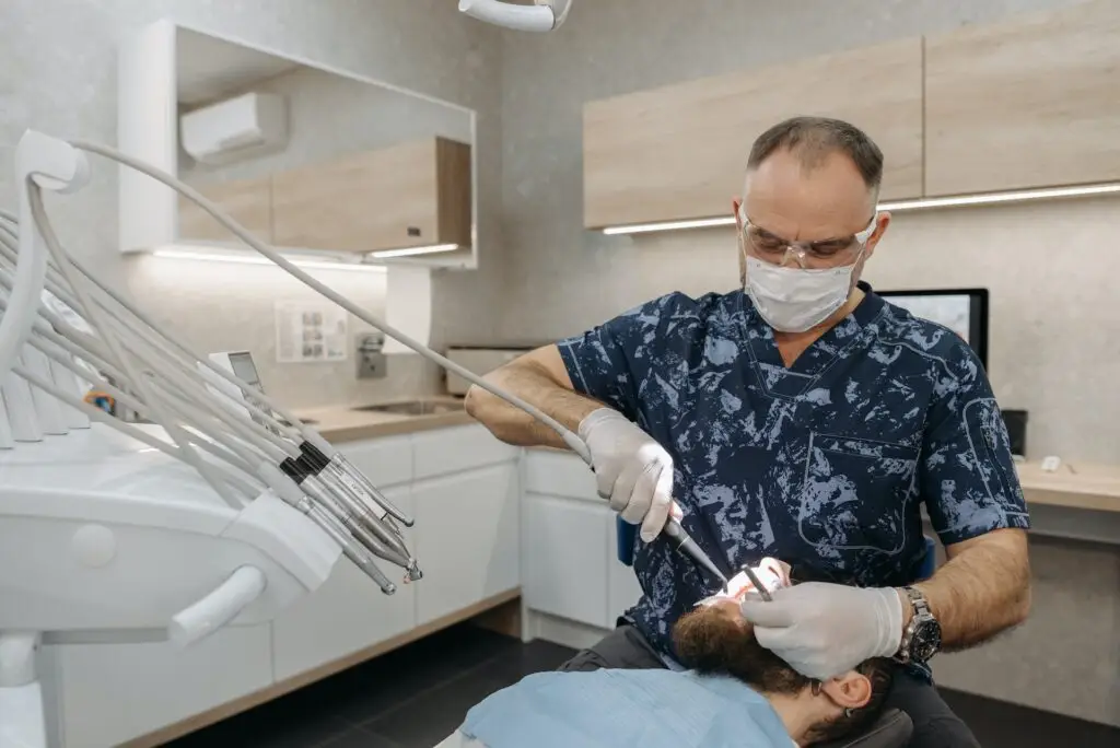 Dentysta w masce, rękawiczkach i okularach ochronnych bada jamę ustną pacjenta w nowoczesnej klinice stomatologicznej. Pacjent, wygodnie leżący na fotelu dentystycznym w jasnym świetle, poddawany jest zabiegowi sedacji wziewnej dla relaksu.