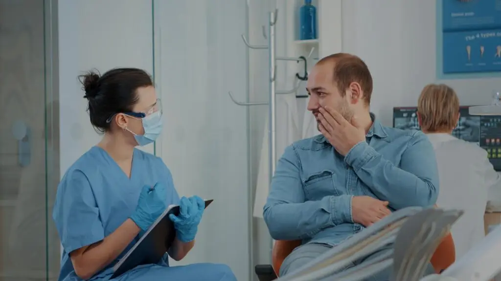 Dentysta w fartuchu i maseczce omawia możliwe leczenie bólu zęba z pacjentem w klinice stomatologicznej, podczas gdy inny członek personelu pracuje przy komputerze w tle.