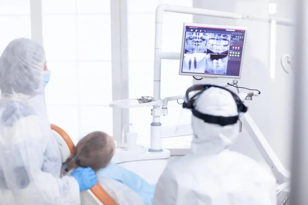 Dwóch dentystów w odzieży ochronnej ogląda zdjęcia rentgenowskie pacjenta wyświetlane na monitorze w jasnej klinice, omawiając opcje leczenia retencyjnego. Pacjent leży wygodnie na fotelu dentystycznym.