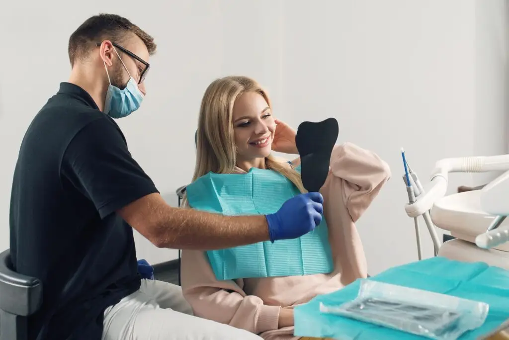Dentysta w masce i rękawiczkach pokazuje uśmiechniętej kobiecie jej odbicie w ręcznym lusterku podczas przeglądu stomatologicznego. Pacjentka siedzi na fotelu dentystycznym ze śliniakiem, omawiając plan leczenia stomatologicznego. Narzędzia dentystyczne znajdują się na tacy obok.