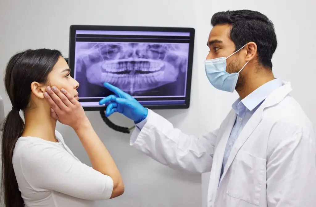 Dentysta w masce i rękawiczkach omawia zdjęcie rentgenowskie zęba pokazujące zatrzymany ząb z pacjentką, która dotyka swojego policzka, w otoczeniu kliniki. Zdjęcie rentgenowskie jest wyświetlane na monitorze za nimi.