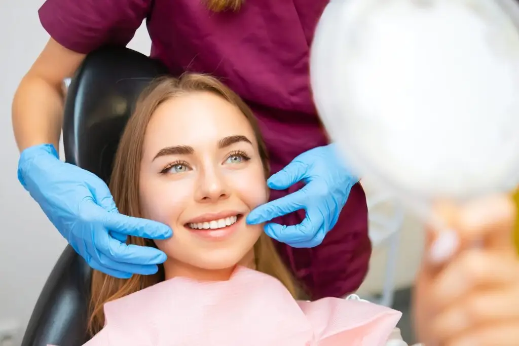 Kobieta siedzi na fotelu dentystycznym, uśmiechając się, podczas gdy dentysta w rękawiczkach bada jej zęby. Trzyma ręczne lusterko i nosi różowy śliniak, prawdopodobnie omawiając zalecenia po wyrwaniu zęba podczas przeglądu dentystycznego.