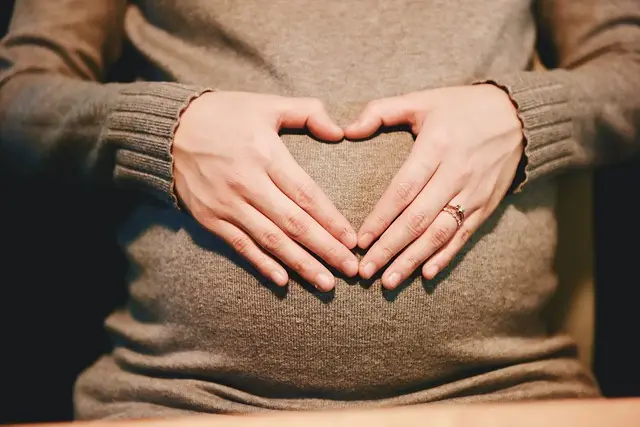 Osoba ubrana w brązowy sweter tworzy dłońmi kształt serca na swoim ciążowym brzuchu, odzwierciedlając chwile radości i pytania takie jak "Czy w ciąży można leczyć zęby", pokazując troskę podjętą podczas ciąży.