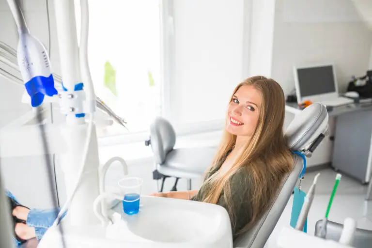 Kobieta z długimi blond włosami siedzi na fotelu dentystycznym, uśmiechając się do kamery po piaskowaniu zębów i skaling. Sprzęt dentystyczny, kubek niebieskiego płynu do płukania ust i komputer są widoczne w jasnym, nowoczesnym gabinecie stomatologicznym.